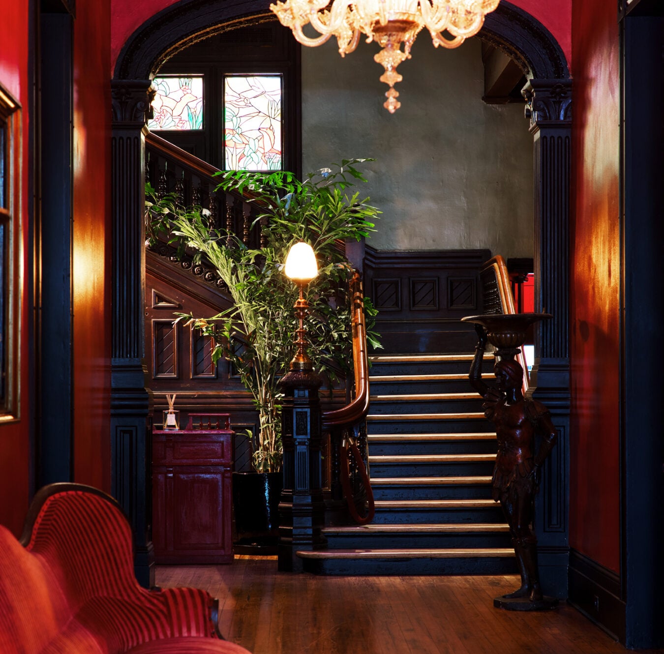 Rich mahogany millwork and stained glass windows frame the grand staircase, where crystal chandeliers cast warm light across polished wood floors and classic Victorian elegance.