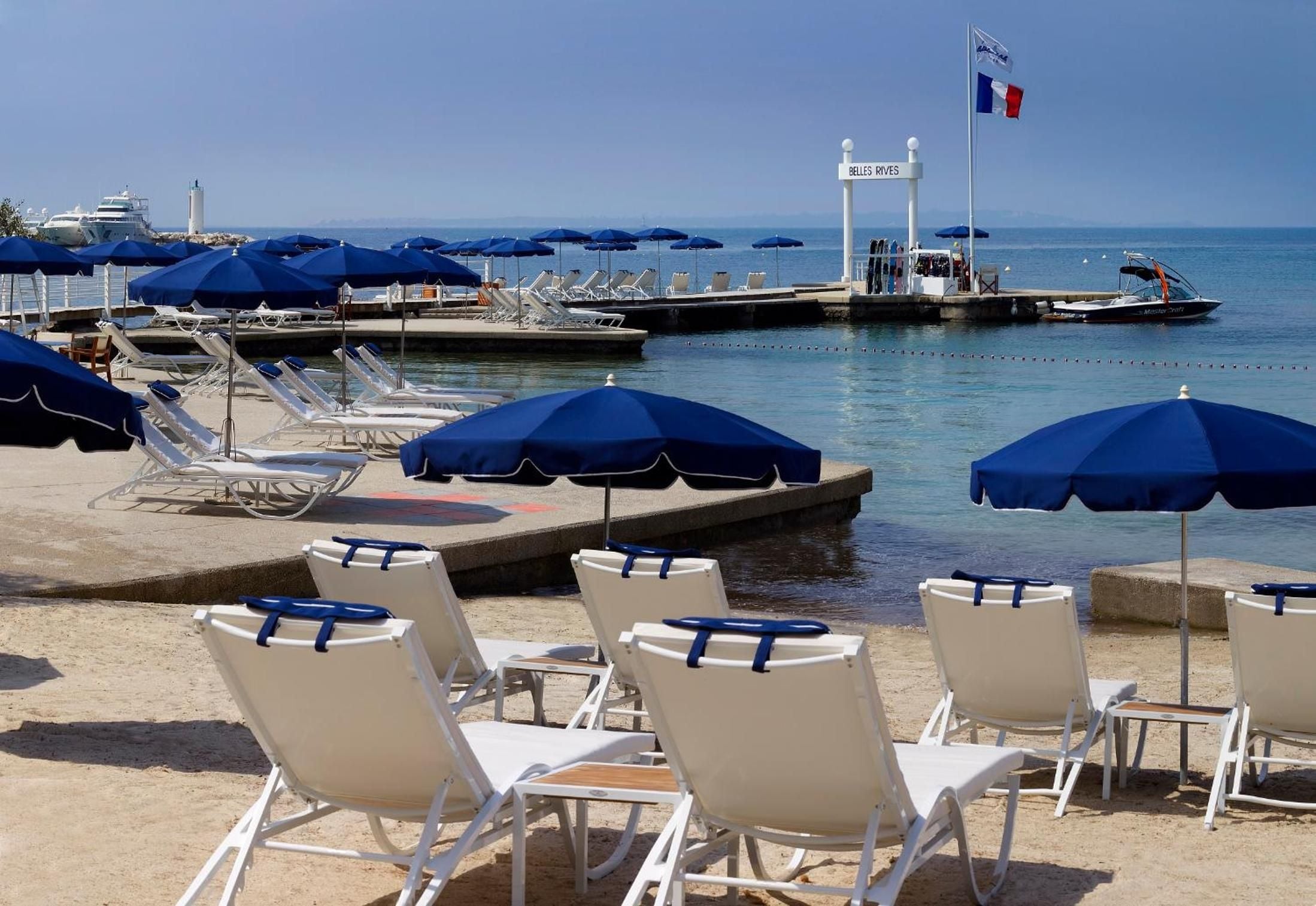 Blue-striped umbrellas and crisp lounge chairs dot the private beach platform, where the French Riviera's crystalline waters lap against weathered concrete jetties under azure skies.