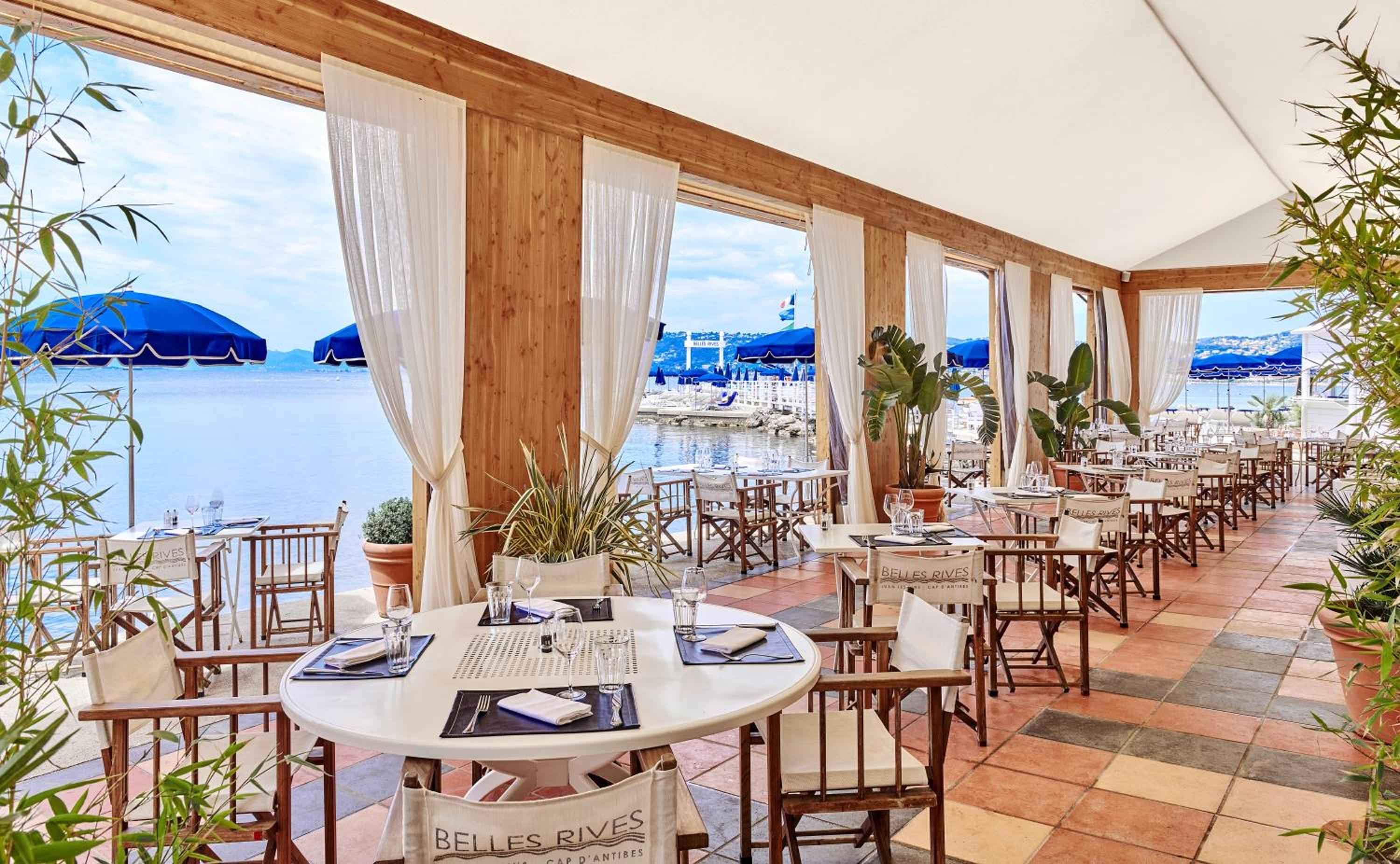 Crisp white linens and bamboo chairs frame azure Mediterranean views at this waterfront terrace, where blue umbrellas dot the shoreline beyond.