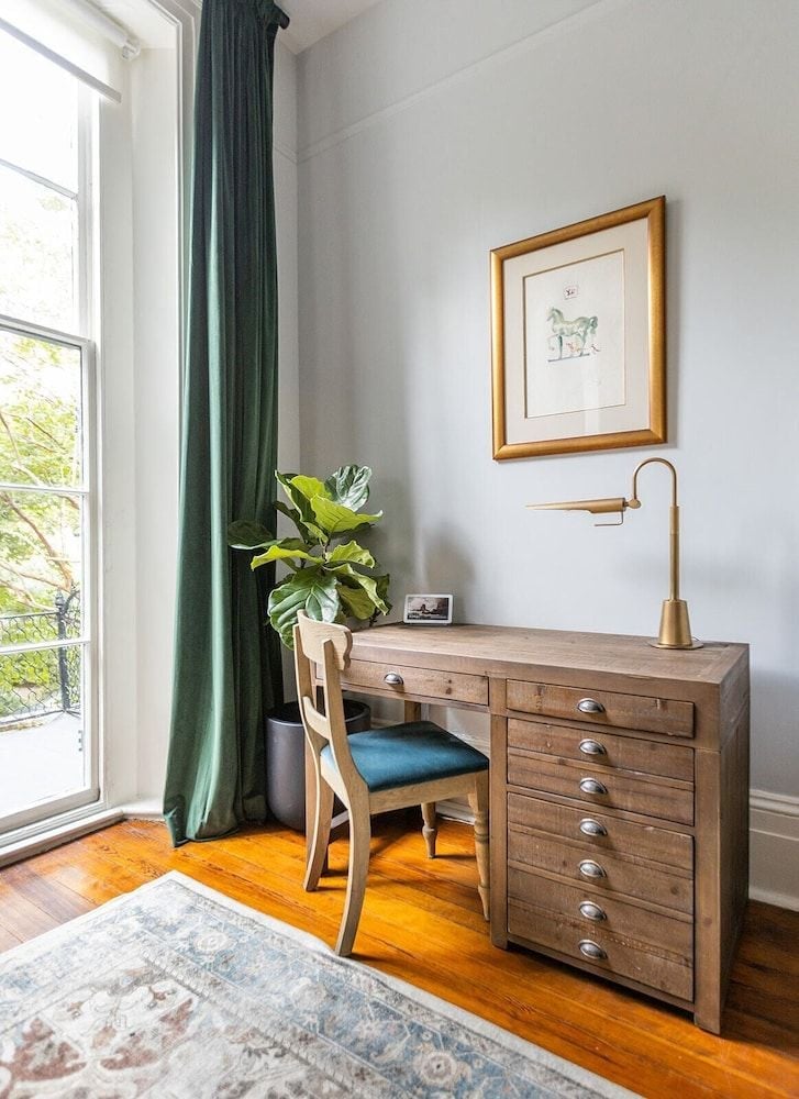 Warm hardwood floors and emerald curtains frame a perfectly appointed writing desk, where brass fixtures and botanical prints create an intimate corner for quiet reflection.