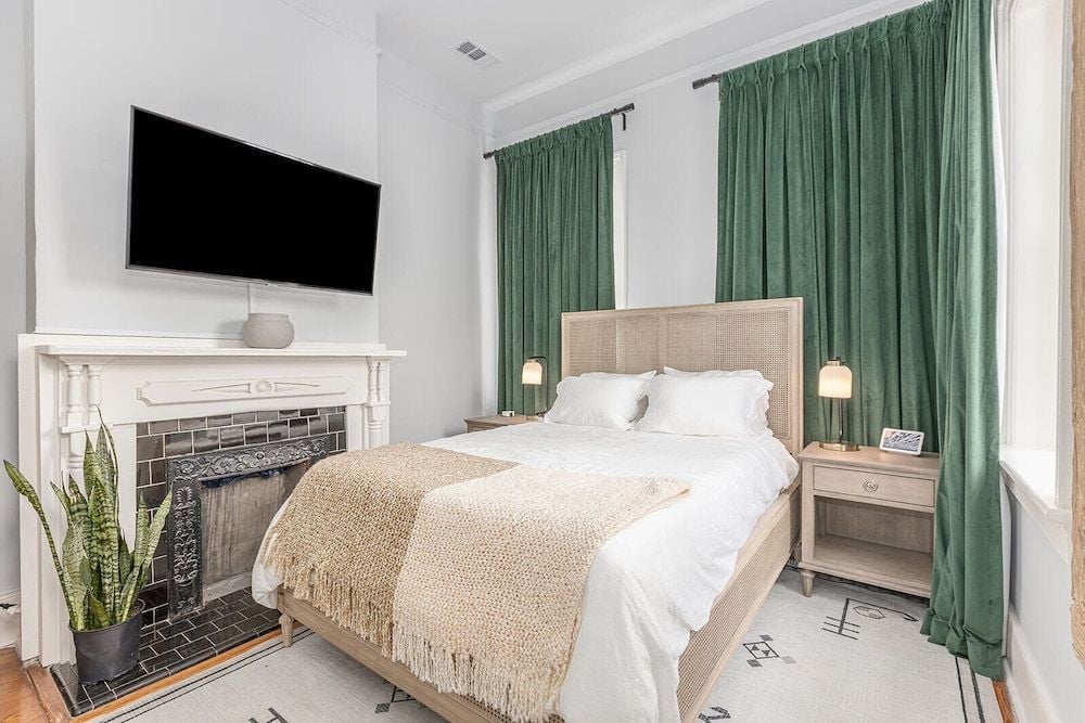 Sage-green curtains frame tall windows while a woven headboard and textured throw create layers of natural warmth against crisp white linens and subway-tiled fireplace.
