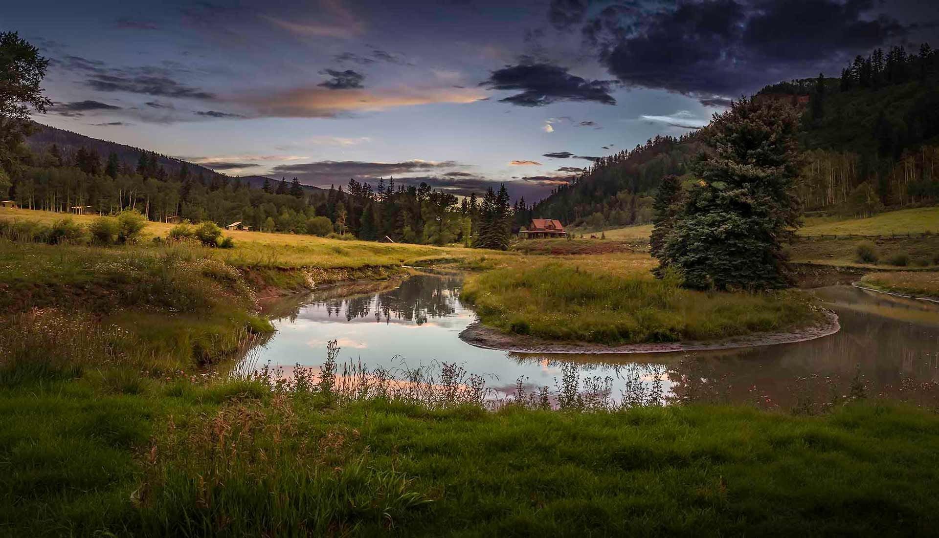 Evening light bathes the meandering river and emerald meadows, where rustic cabins nestle between forested hills under Colorado's dramatic twilight sky.