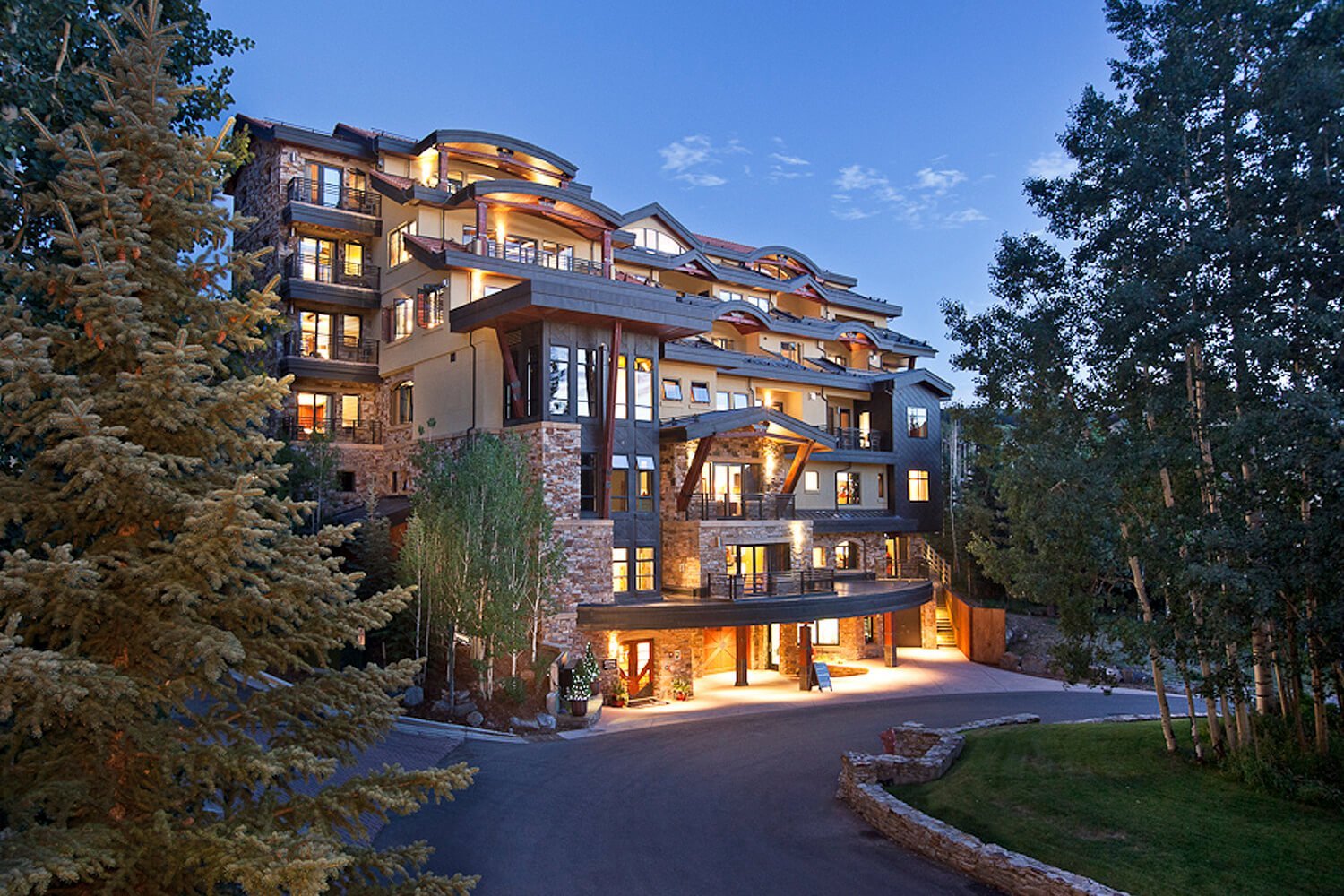 Stone and timber façades cascade down the mountainside, their warm windows glowing against twilight skies as the resort's dramatic architecture emerges from aspen groves.