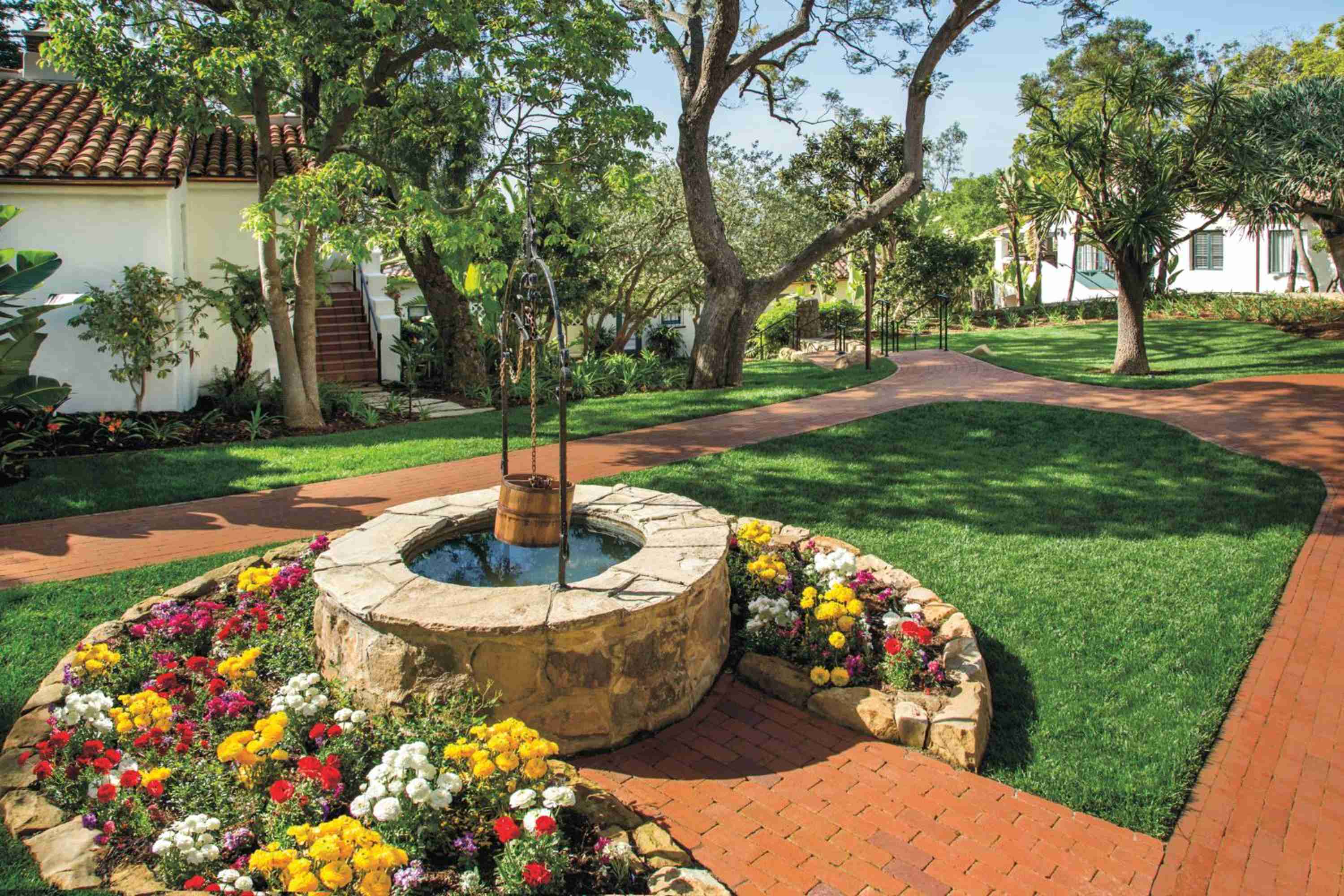 Stone fountains bubble peacefully amid vibrant flower beds, where brick pathways wind through manicured lawns beneath towering eucalyptus trees and Spanish colonial casitas.