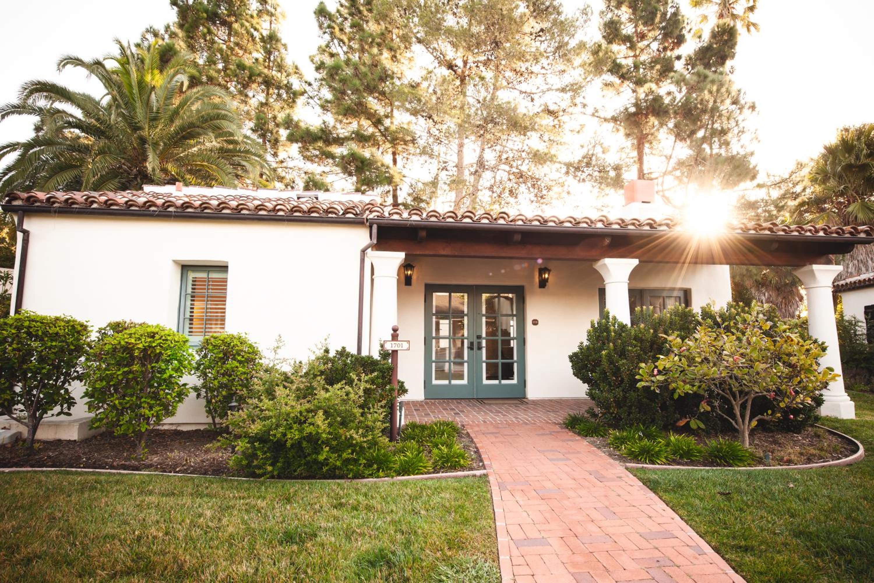 Golden sunlight filters through mature palms and pines, illuminating the Spanish Colonial entrance where terra cotta tiles and sage-green French doors whisper of old California elegance.
