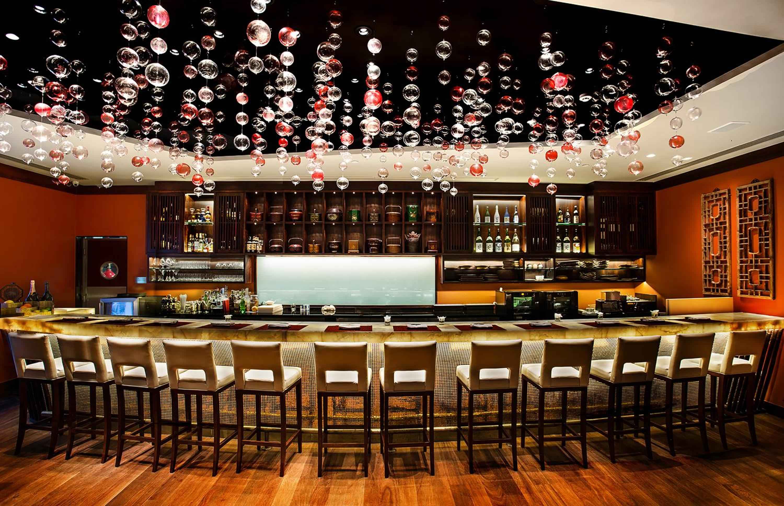 Suspended glass orbs catch amber light above the sleek bar counter, where cognac-hued walls and warm wood create an intimate setting for evening cocktails.