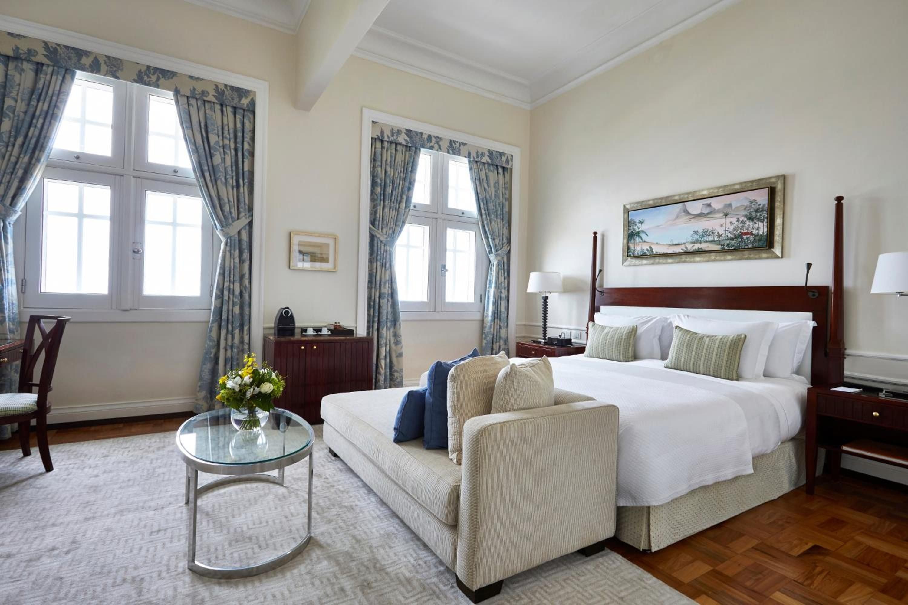 Elegant four-poster bed anchors this refined suite, where blue-patterned curtains frame tall windows and rich mahogany furnishings create timeless Brazilian sophistication.