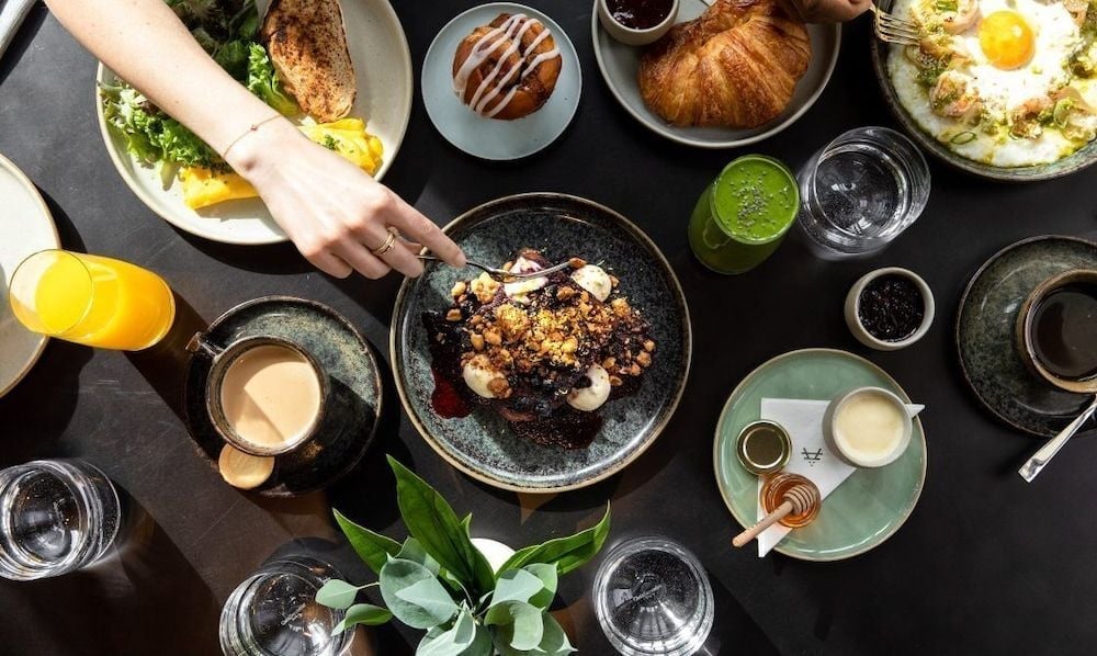 Artfully plated dishes arrive at rustic ceramic tables, where hands reach for coffee-glazed desserts and golden croissants amid morning light and verdant herbs.