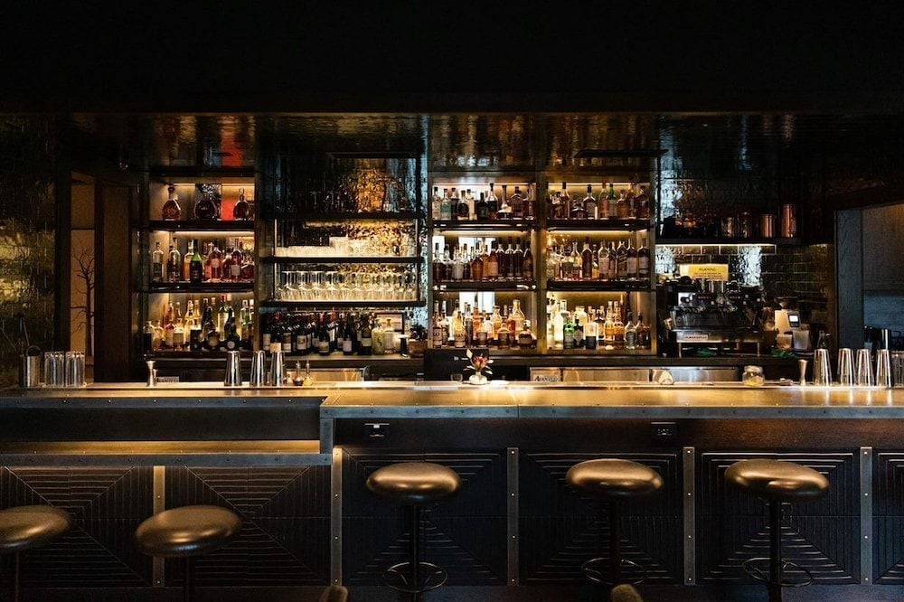 Warm amber light illuminates rows of artisanal spirits and craft cocktails behind polished copper bar stools, creating an intimate speakeasy atmosphere perfect for evening conversations.