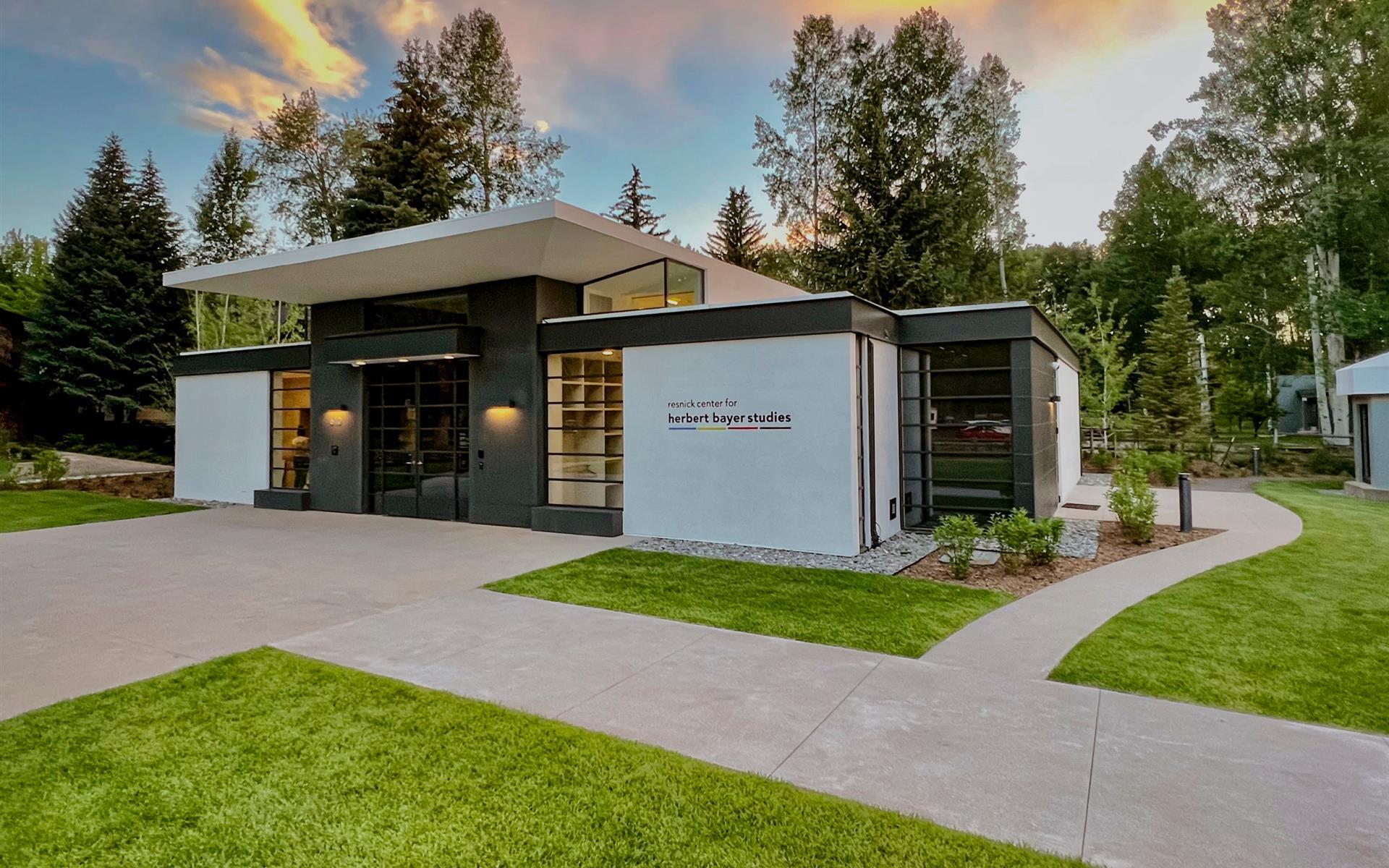 Modern glass and steel lines of the Herbert Bayer Studies center emerge from manicured lawns, where golden hour light catches contemporary architecture against Aspen's towering evergreens.