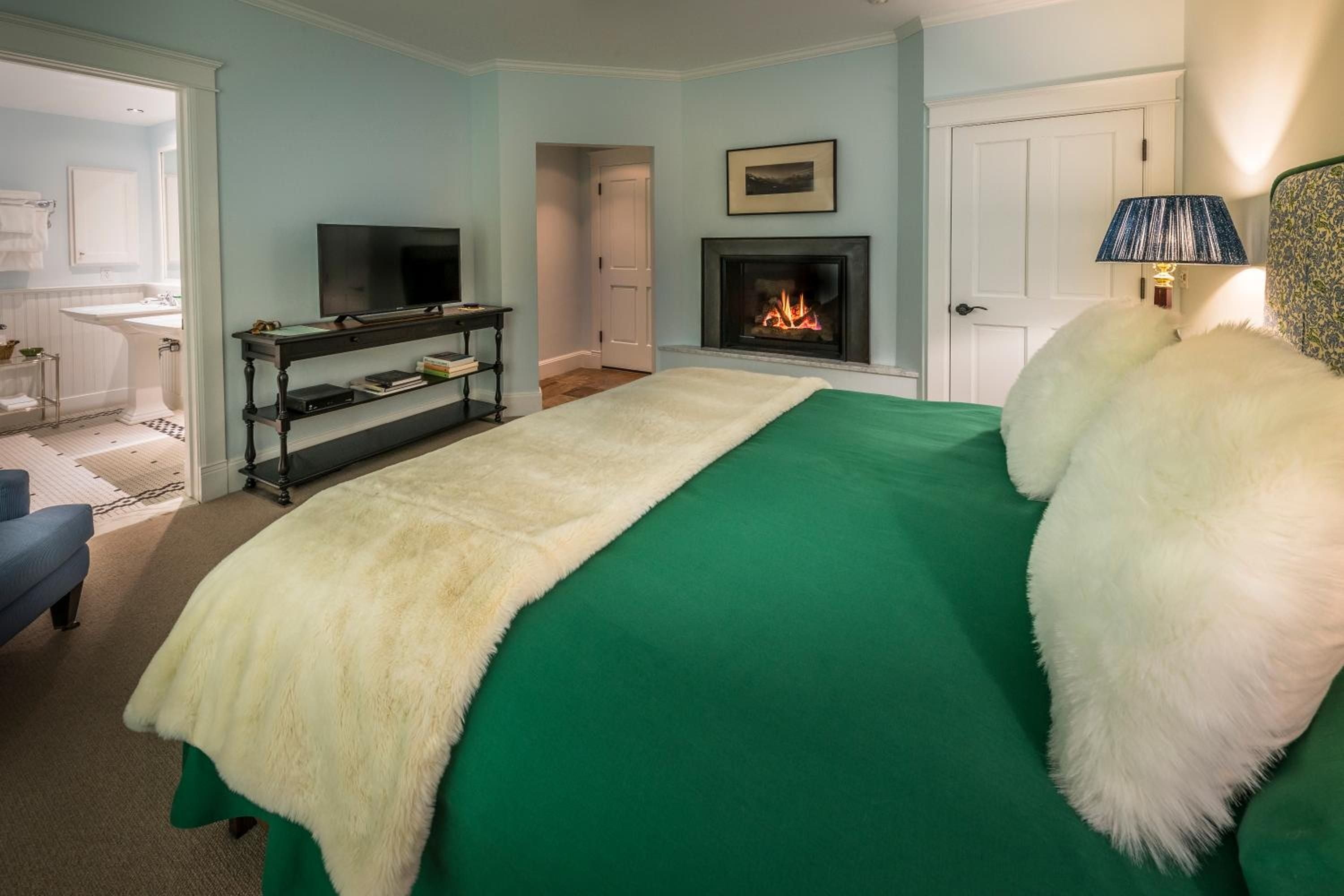 Emerald green linens and plush sheepskin throws create a cozy sanctuary, while the crackling fireplace and soft lamplight cast an intimate glow across this sophisticated bedroom retreat.