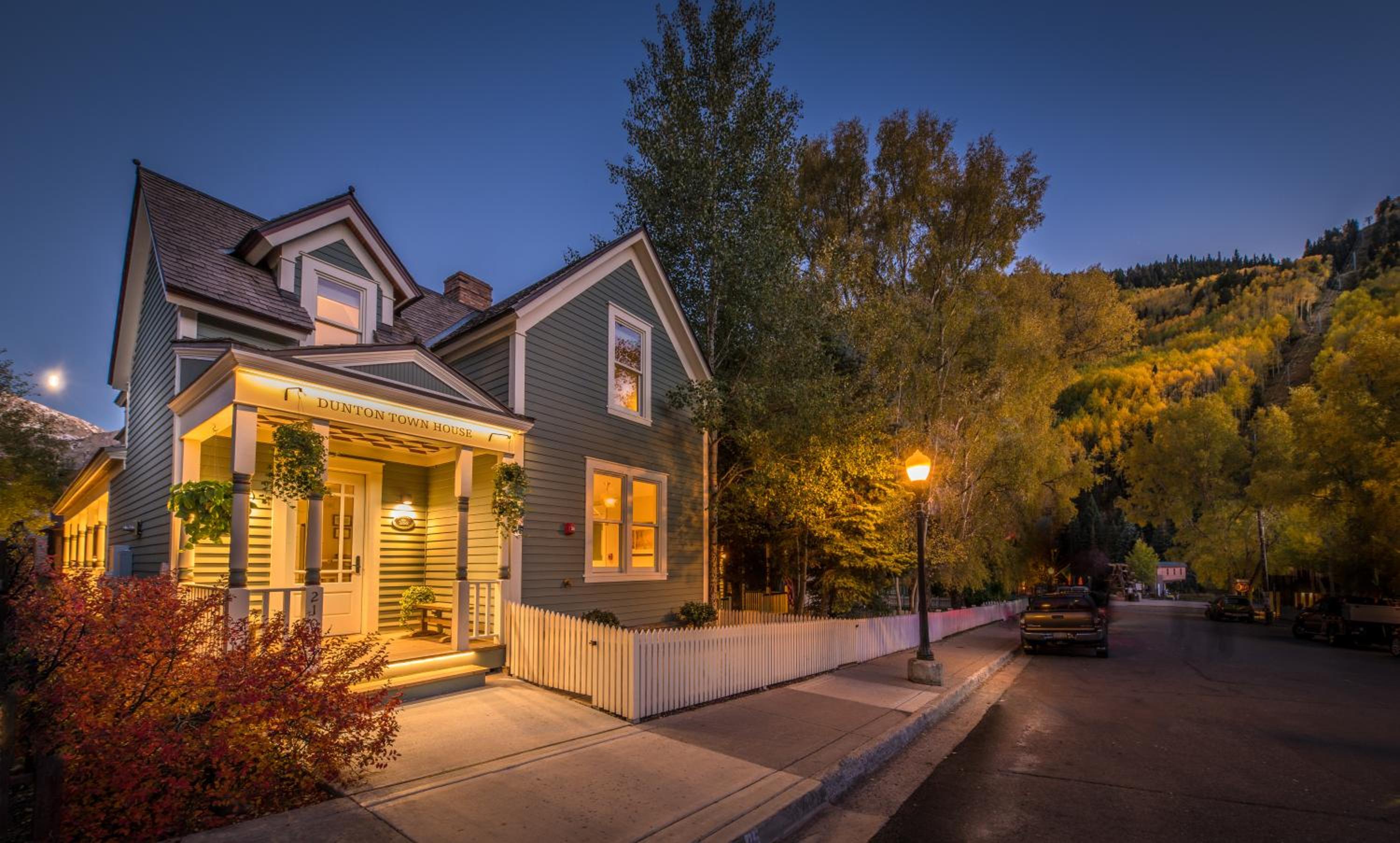 Warm light spills from the Victorian-era windows of this intimate mountain retreat, where autumn aspens frame the charming clapboard facade against twilight skies.