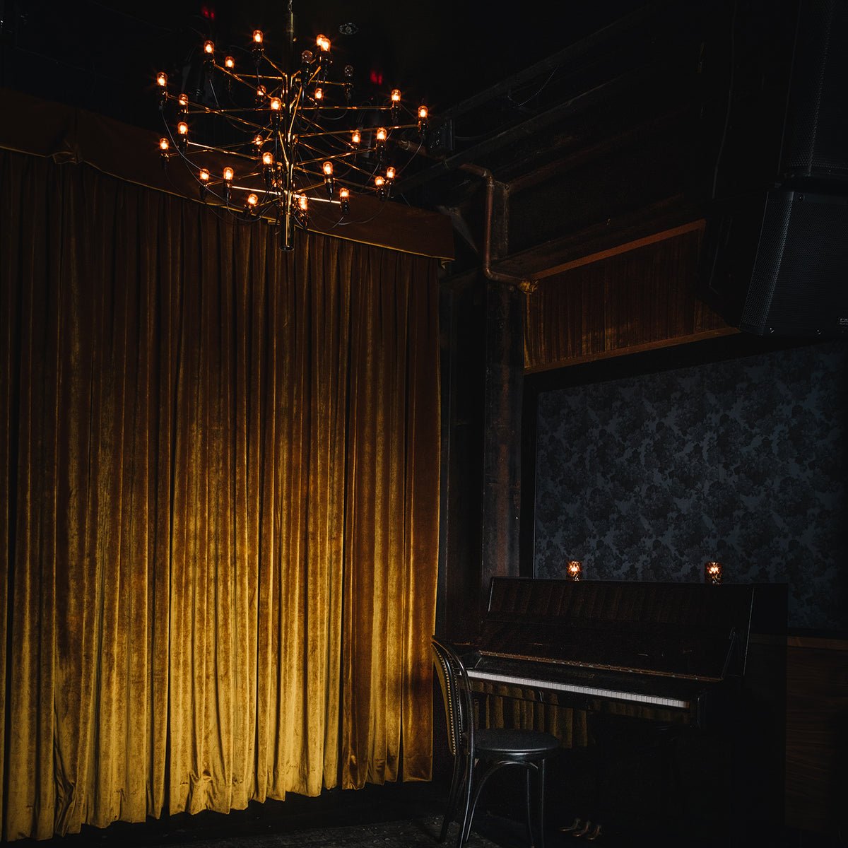 Amber velvet curtains and industrial chandeliers create an intimate speakeasy atmosphere where vintage charm meets modern edge in this moody corner nook.