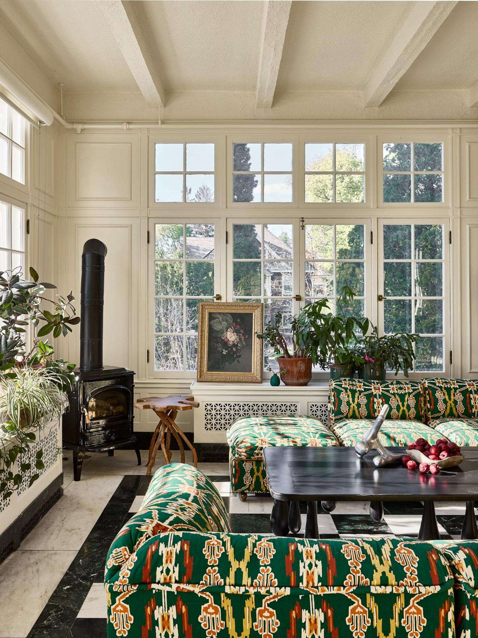 Sunlight streams through divided-light windows onto richly patterned green textiles and polished wood, creating an intimate gathering space warmed by the cast-iron stove.