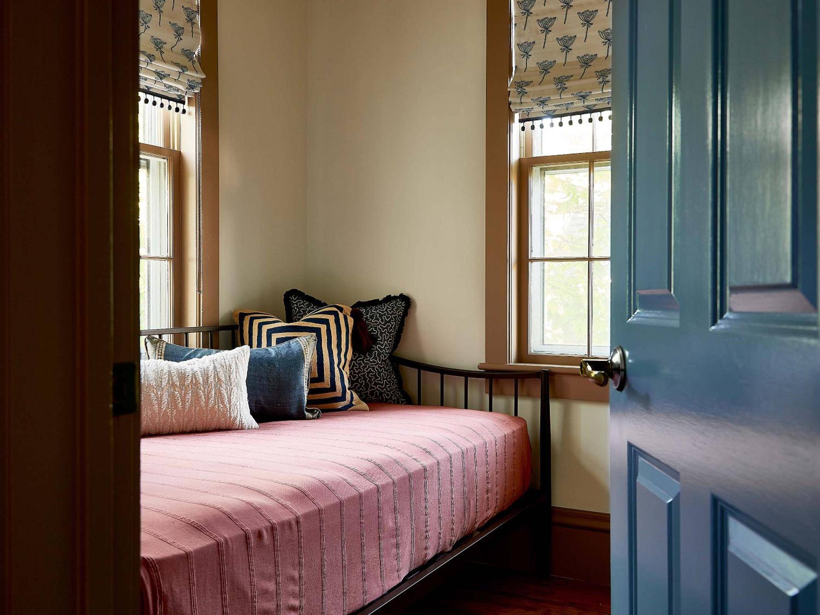 Natural light filters through patterned Roman shades onto a coral-pink daybed adorned with geometric pillows, creating an intimate reading nook beside weathered wood windows.