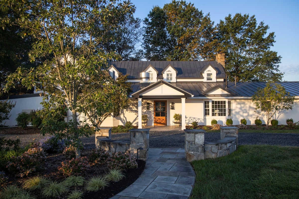 Stone pathways wind through thoughtfully landscaped gardens toward Blue Rock's welcoming covered entrance, where dormer windows and warm lighting create an intimate country estate atmosphere.