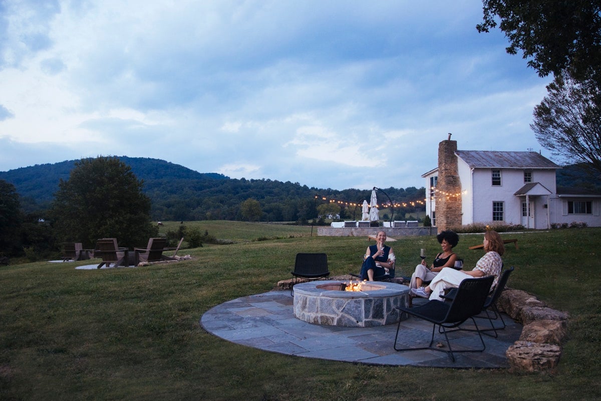 Guests gather around the stone fire pit as twilight settles over rolling hills, string lights casting a warm glow across the pastoral landscape.