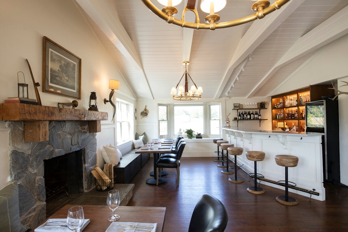 Rustic wooden beams and stone fireplace create intimate dining quarters, while the marble bar beckons with golden lighting and artfully arranged spirits.