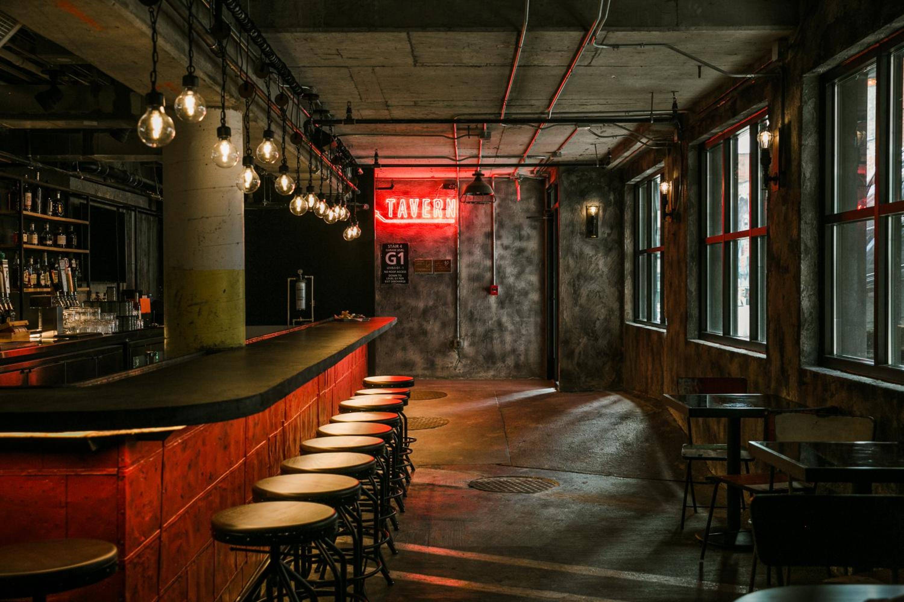 Edison bulbs cast warm amber light across weathered concrete and exposed brick, while the glowing red TAVERN sign beckons guests into this moody industrial-chic watering hole.