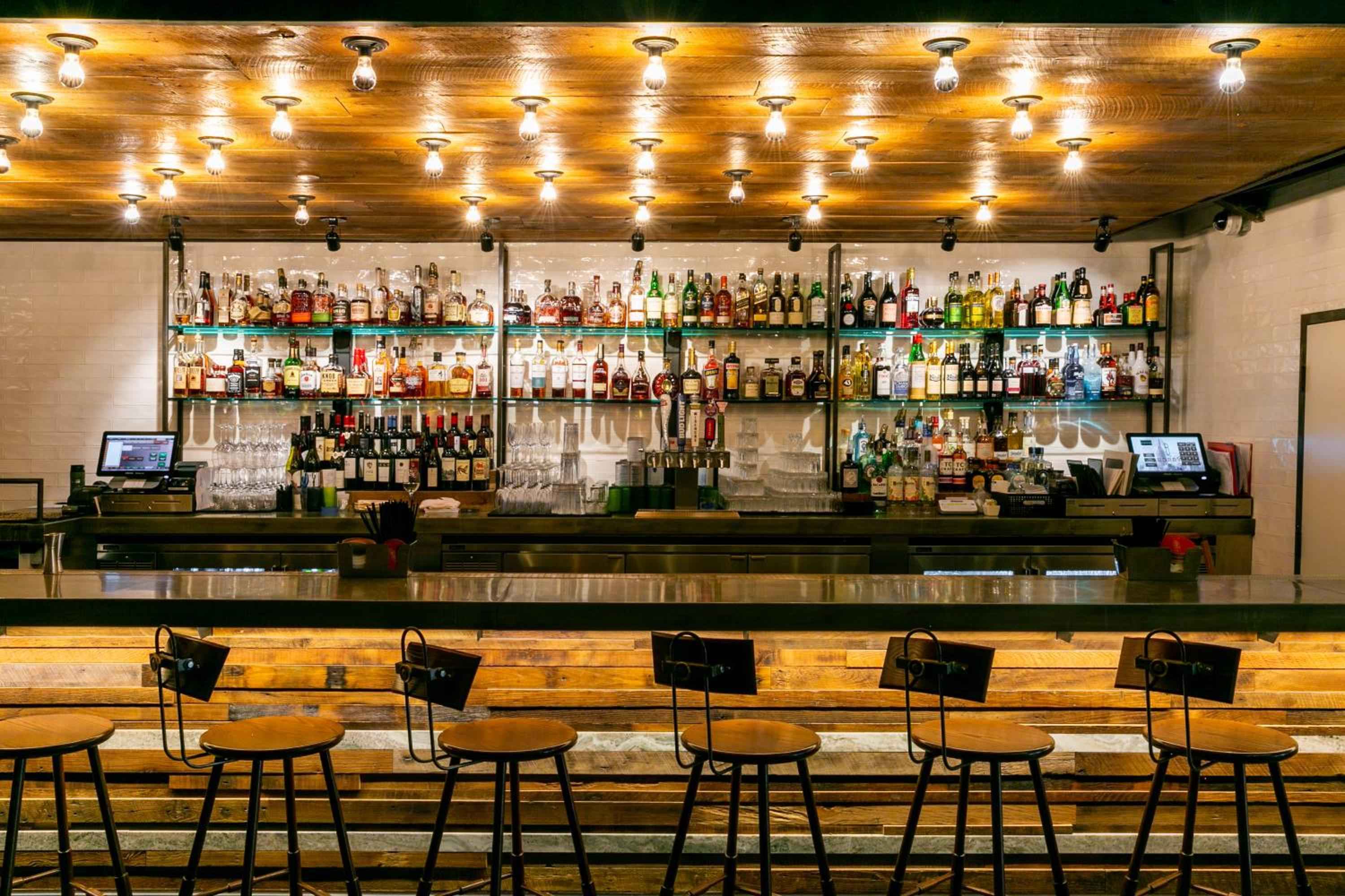 Warm Edison bulbs cast an amber glow across reclaimed wood while hundreds of spirits line industrial shelving, creating an inviting neighborhood bar atmosphere with modern edge.