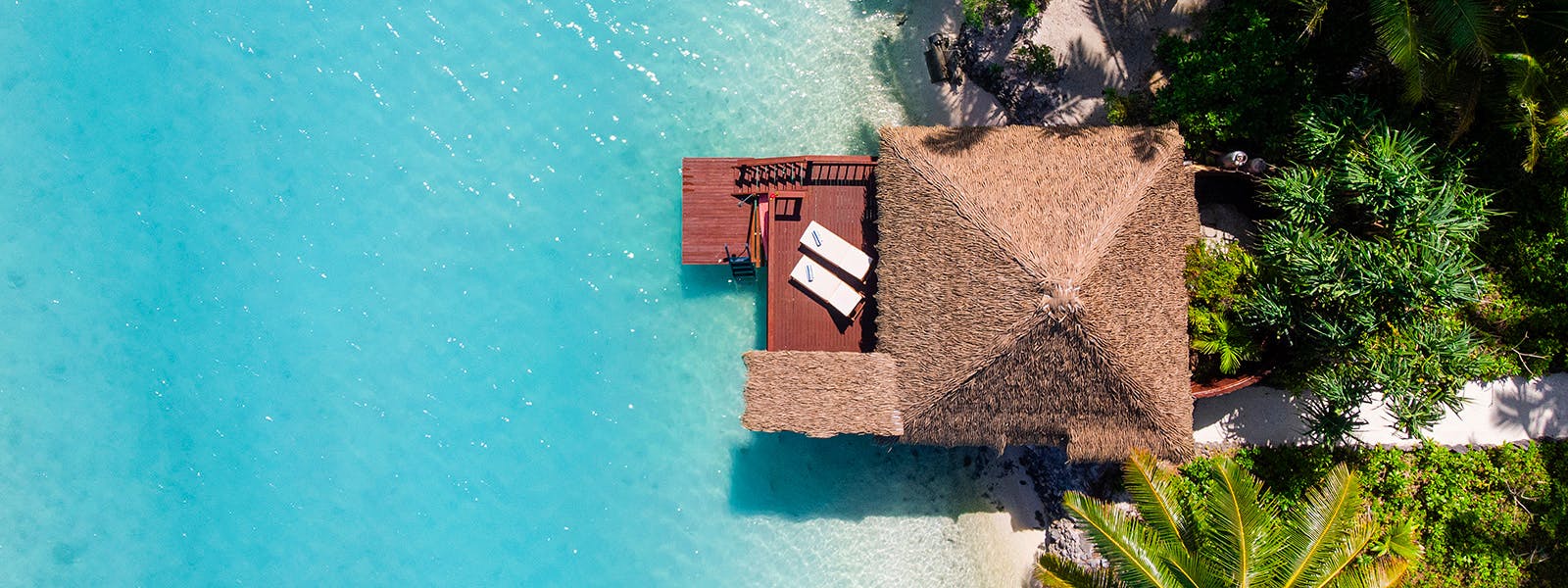 Overwater bungalows with traditional thatched roofs stretch into crystalline lagoon waters, where powder-soft sand meets lush tropical palms in perfect island harmony.