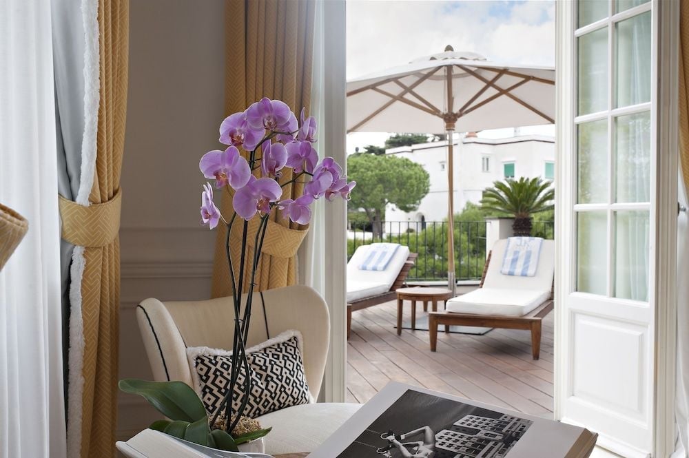 Purple orchids grace a sun-drenched suite where golden curtains frame terraced views and polished wood floors lead to loungers beneath cream umbrellas.
