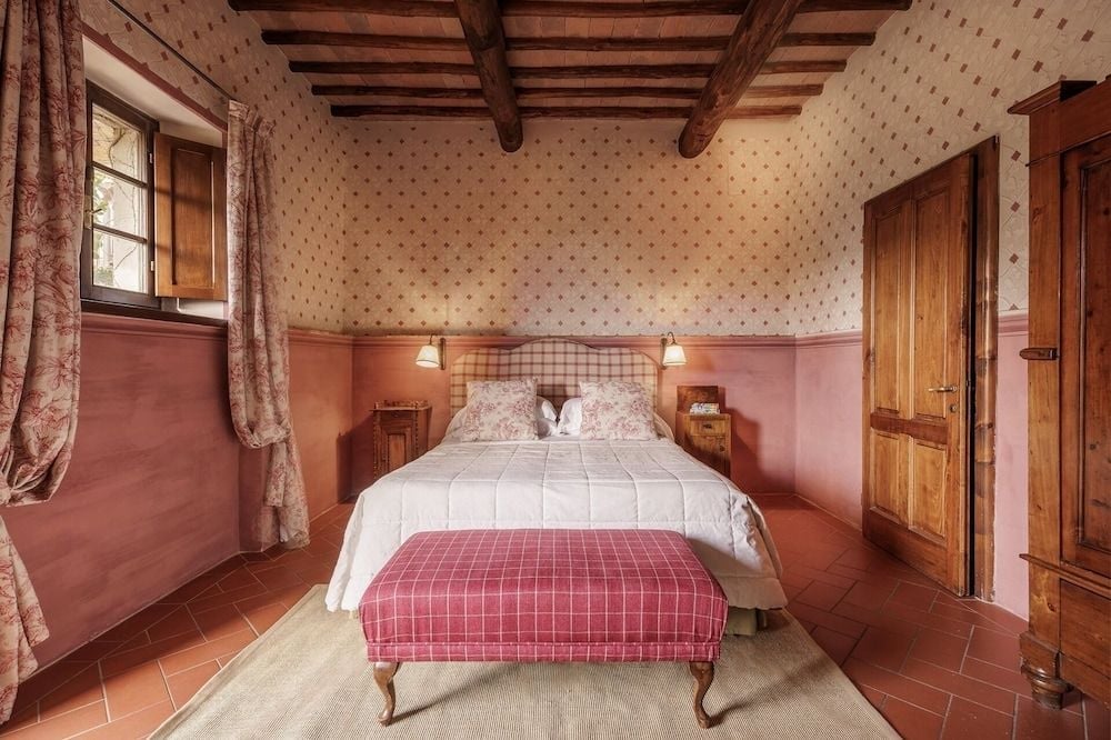Exposed chestnut beams frame this intimate guest room where terracotta floors meet rose-hued walls, creating a quintessentially Tuscan sanctuary filled with rustic charm.