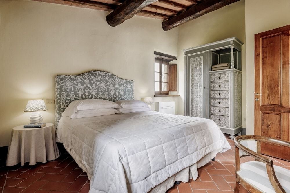 Exposed timber beams frame this intimate guest room where sage-green armoire meets terra cotta floors, creating rustic Tuscan elegance beneath honey-colored ceiling rafters.