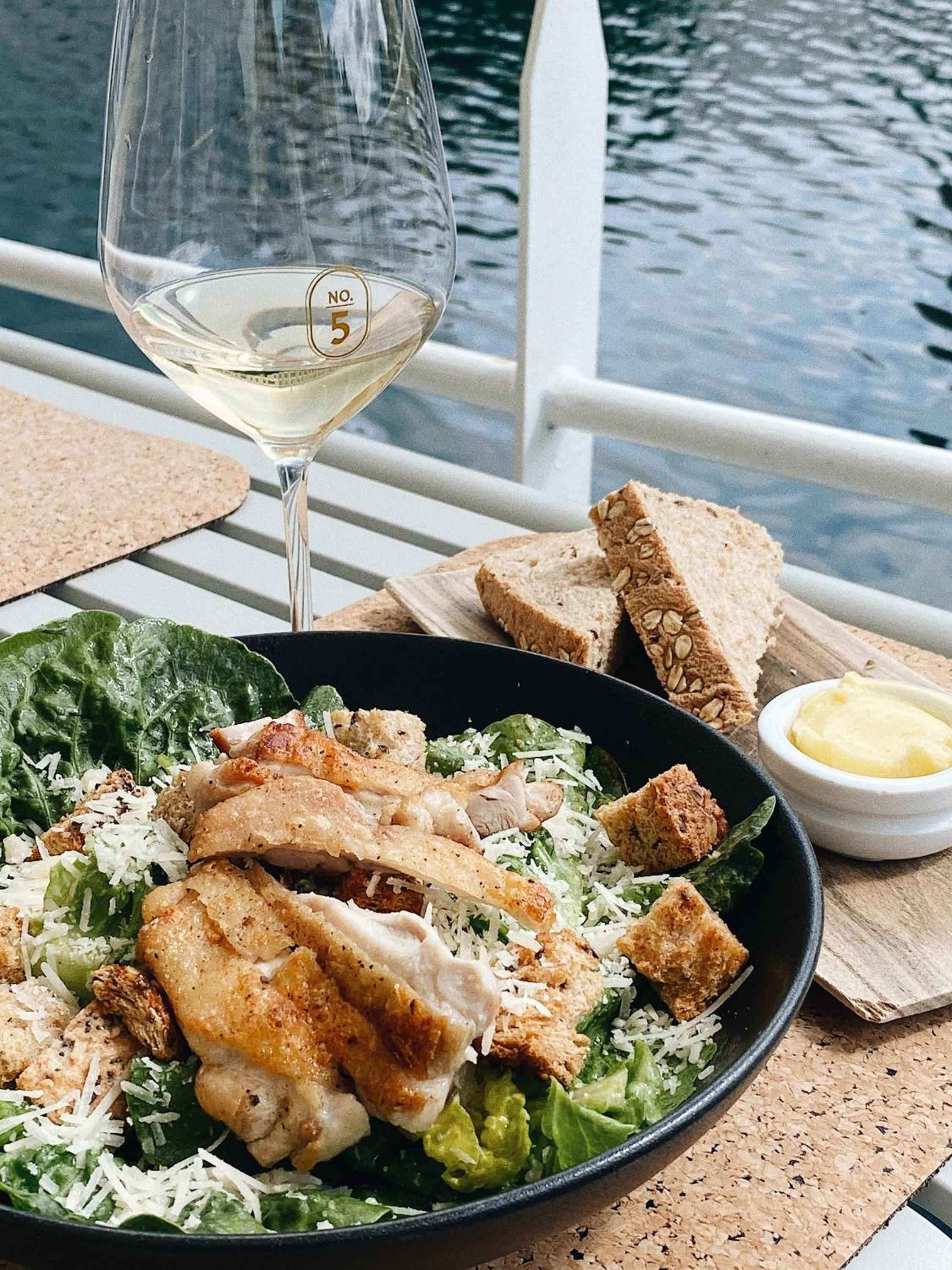 Waterside dining perfection: crisp Caesar salad with golden chicken, crusty artisan bread, and chilled white wine overlooking rippling harbor waters from the hotel's restaurant terrace.