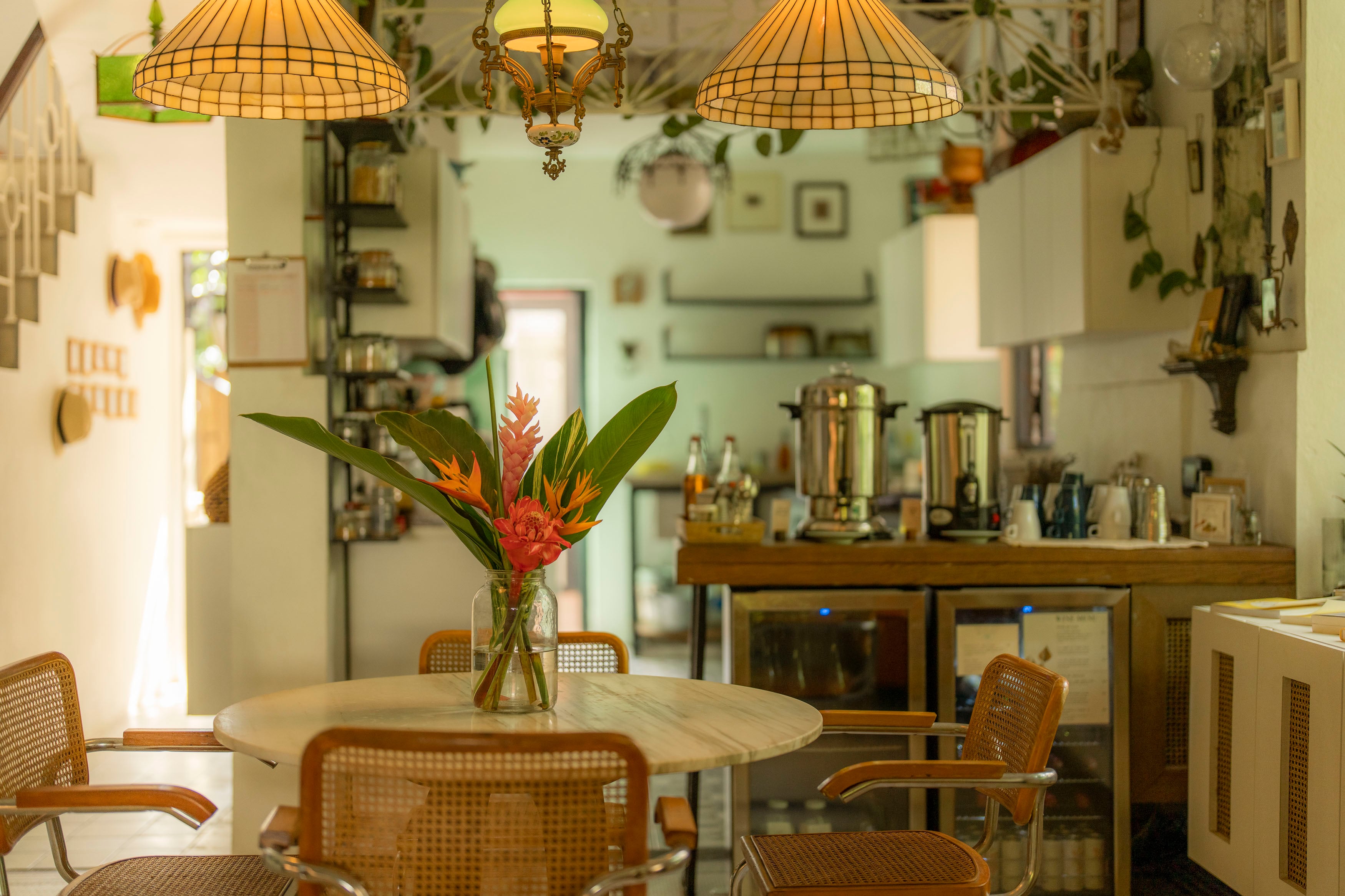 Tropical blooms brighten the airy dining space where woven cane chairs gather beneath warm Tiffany-style pendant lights and polished copper coffee urns gleam.