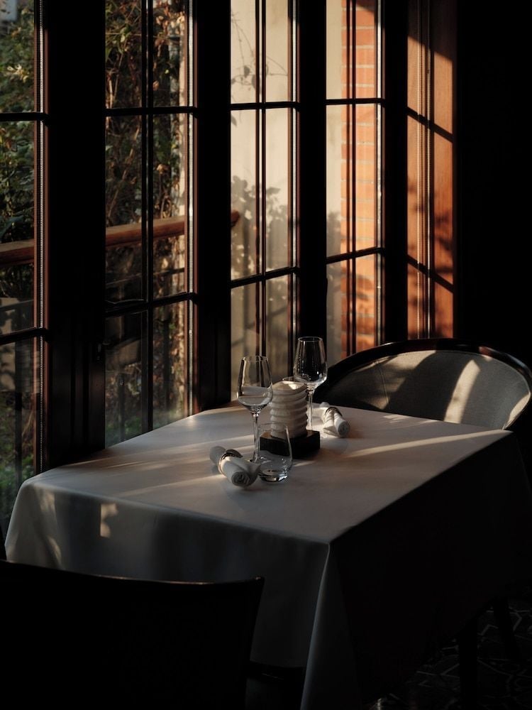 Golden hour light filters through steel-framed windows, casting dramatic shadows across crisp white linens and gleaming glassware in the hotel's intimate dining space.