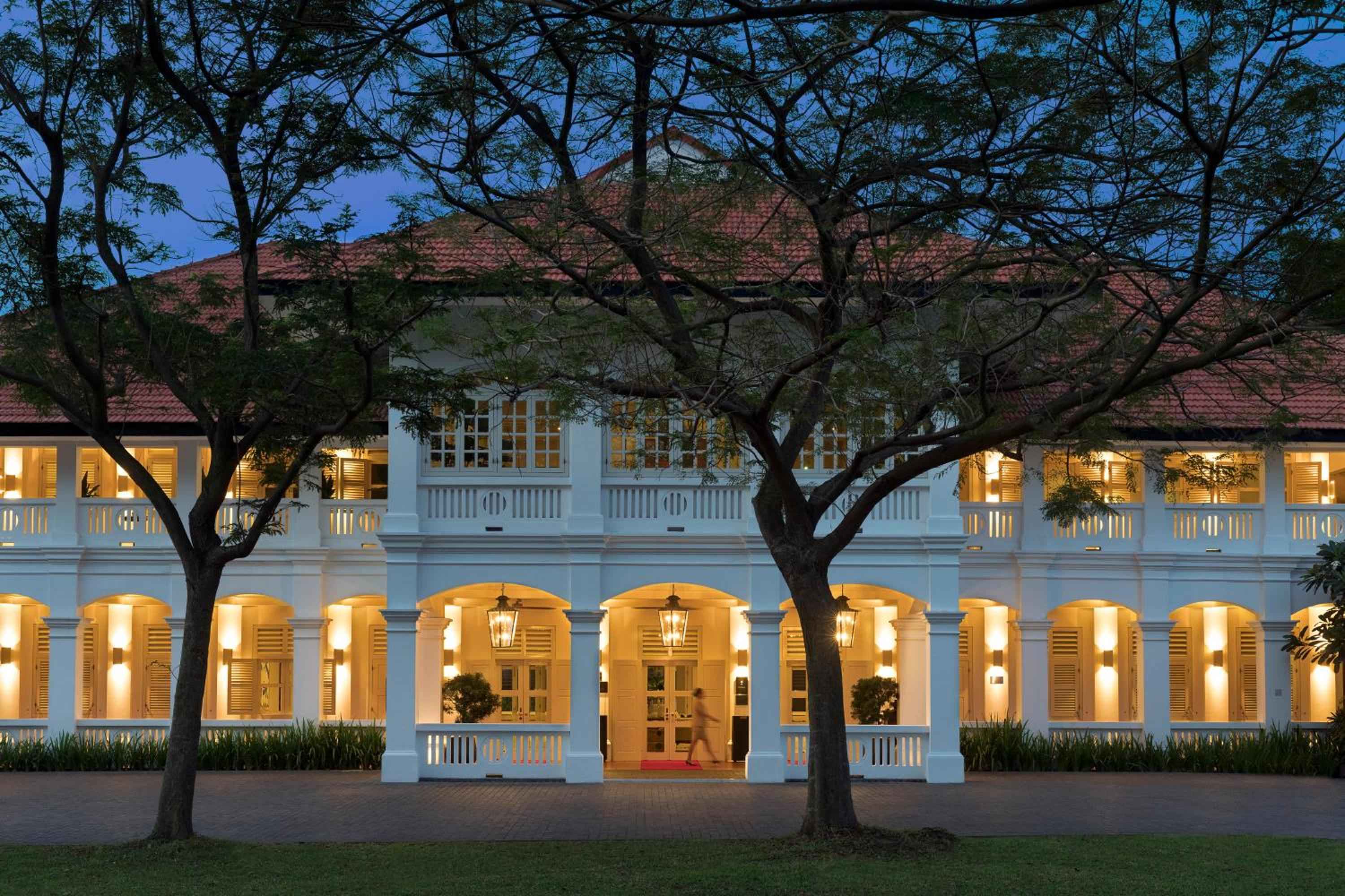 Elegant colonial arches glow warmly beneath a terracotta roof, while towering rain trees frame this sophisticated entrance like natural cathedral pillars at dusk.