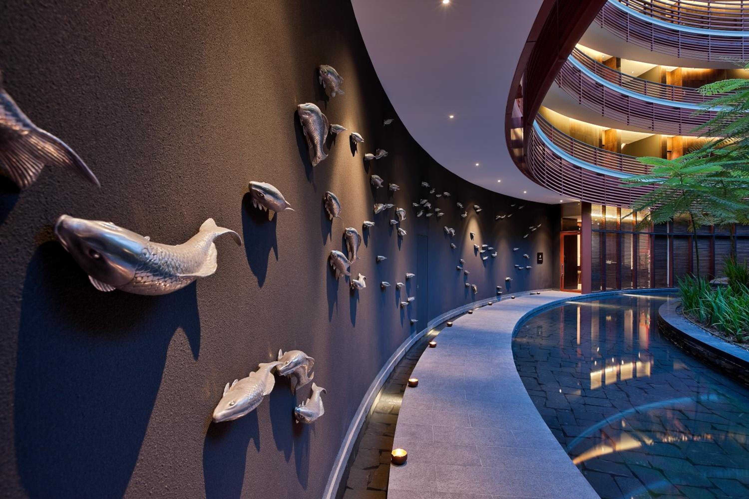 Sculptural fish installations cascade down curved walls above the indoor pool, where warm lighting and flowing water create an aquatic dreamscape beneath soaring contemporary architecture.
