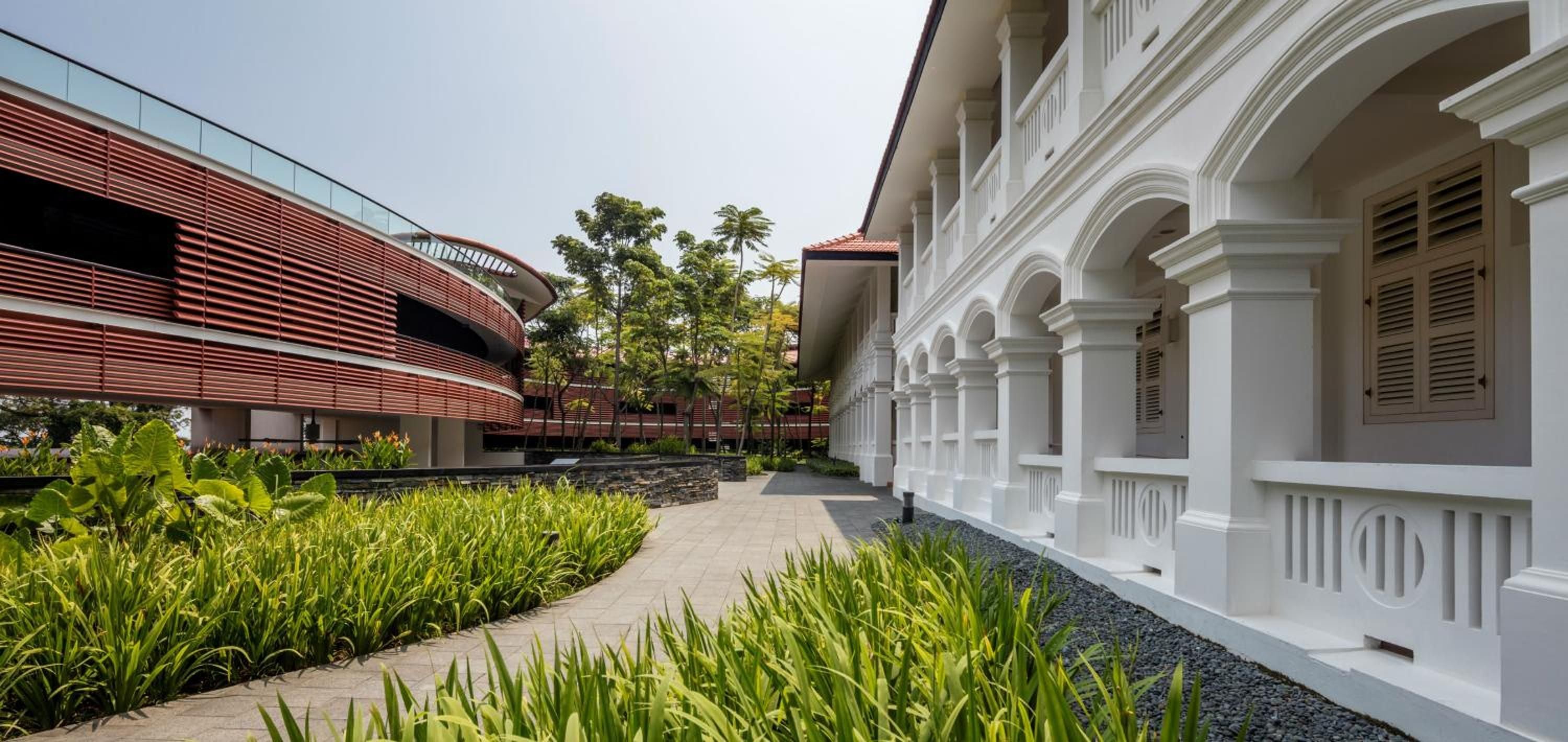 Colonial white arches frame manicured walkways where contemporary red timber structures blend seamlessly with heritage design, creating an intimate courtyard sanctuary amid tropical greenery.