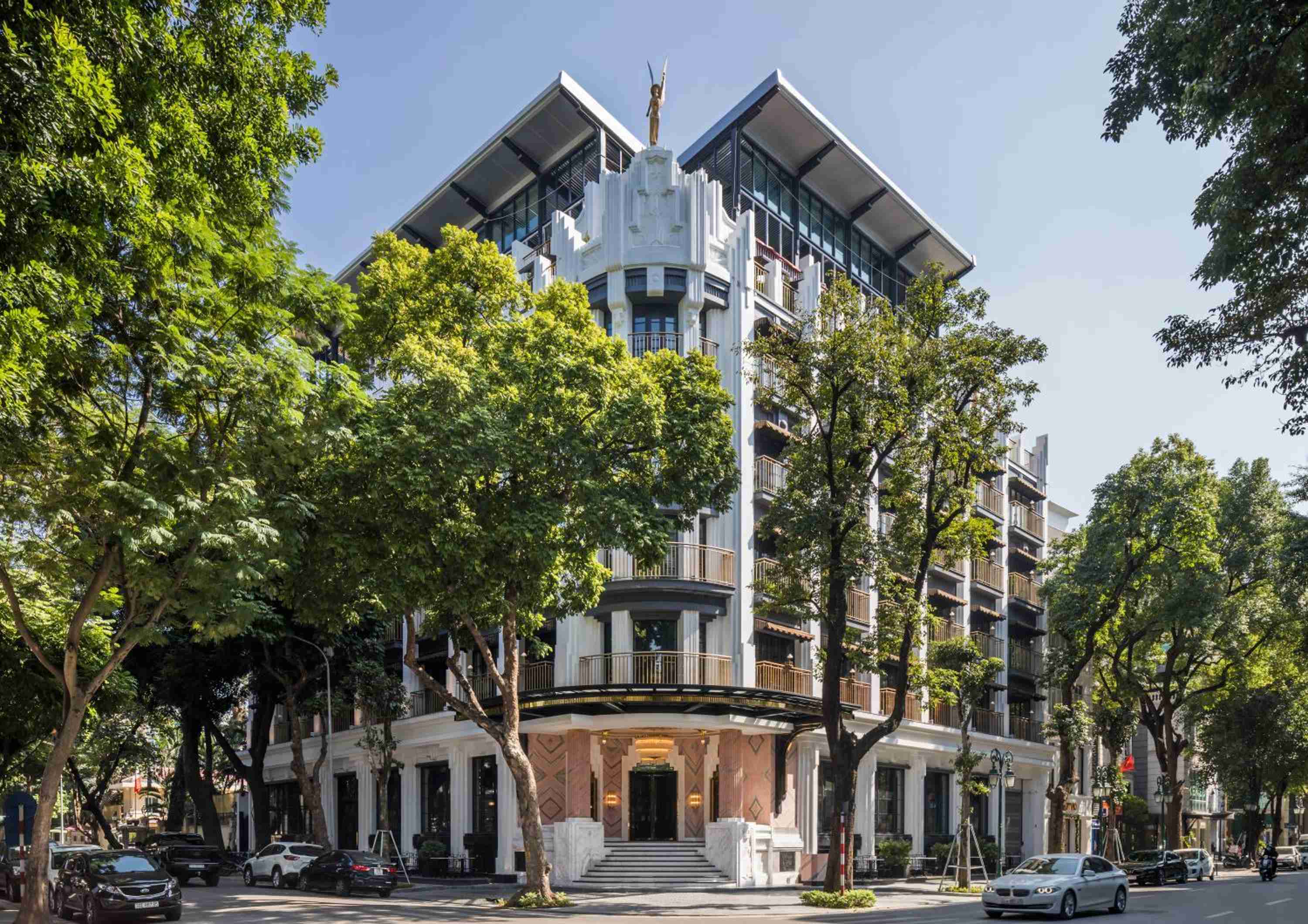 Art Deco elegance meets contemporary Vietnamese design as Capella Hanoi's curved facade rises gracefully through leafy canopy, its warm stone details catching golden hour light.