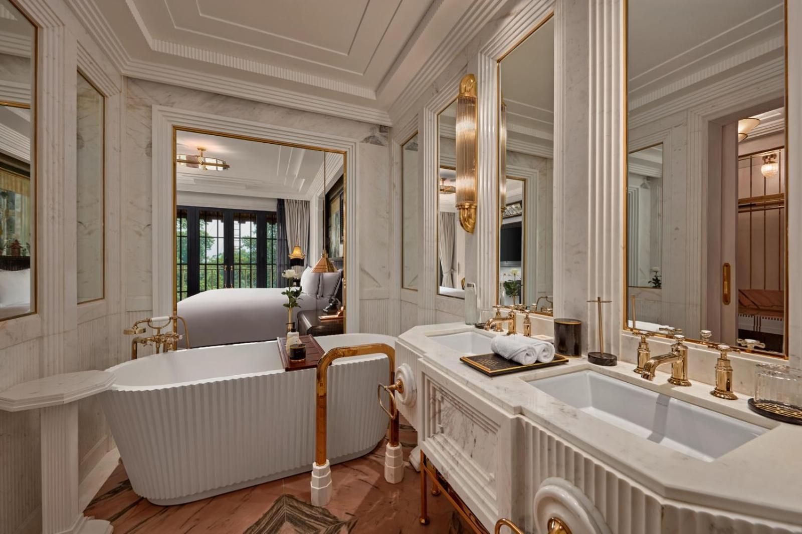 Marble surfaces gleam under golden sconces while a freestanding tub overlooks the bedroom, creating an intimate sanctuary where French colonial elegance meets modern luxury.
