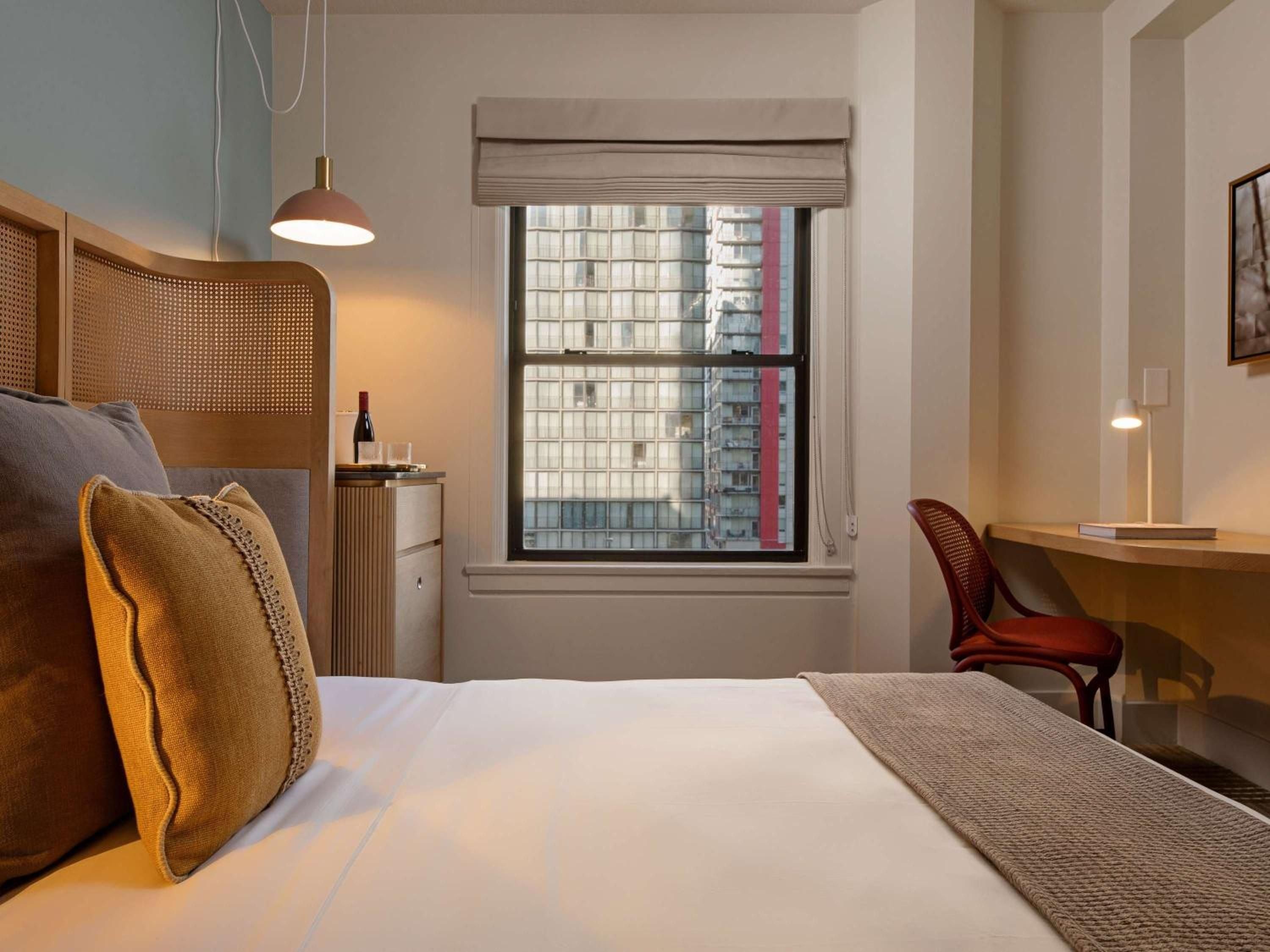 Warm cane headboards and copper pendant lighting create intimate ambiance in thoughtfully appointed guest rooms, where urban views frame a cozy workspace.
