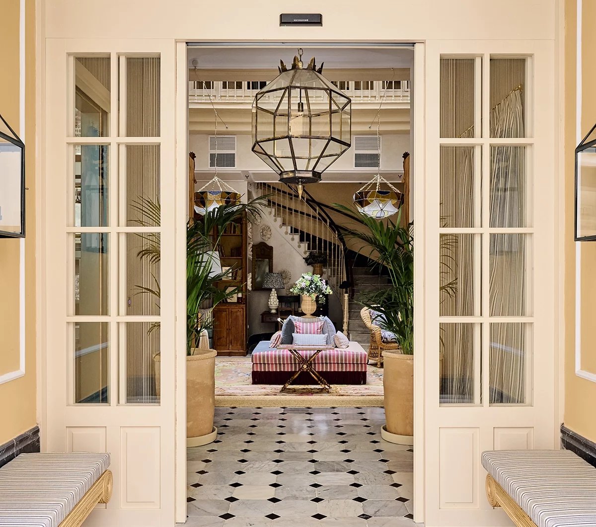 Geometric pendant lights cast warm shadows across checkered marble floors, while French doors frame intimate seating areas adorned with tropical palms and colonial charm.