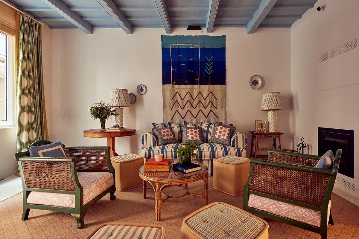 Exposed beams frame a cozy sitting area where striped sofas and woven chairs invite intimate conversations beneath vibrant tapestries and golden afternoon light.