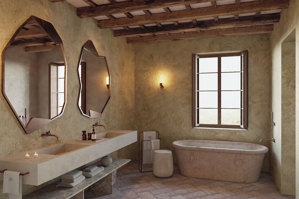Exposed wooden beams frame this serene stone bathroom where geometric mirrors reflect candlelight and a sculptural soaking tub invites contemplation beneath timeless Mediterranean architecture.