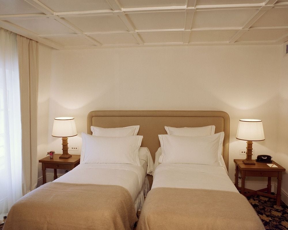 Twin beds dressed in crisp white linens anchor this serene suite, where coffered ceilings and warm wood nightstands create an atmosphere of understated elegance.