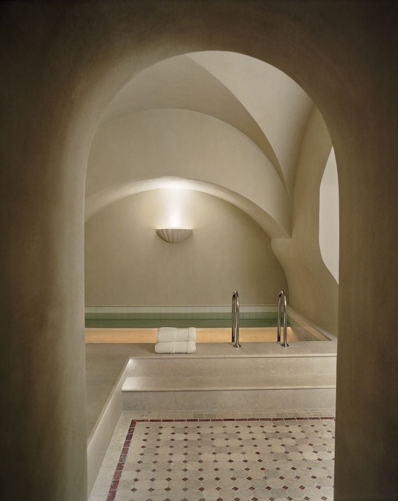Sculptural arches frame the hammam's serene bathing area, where warm light dances across cream-colored stone and geometric mosaic tiles create intimate luxury.