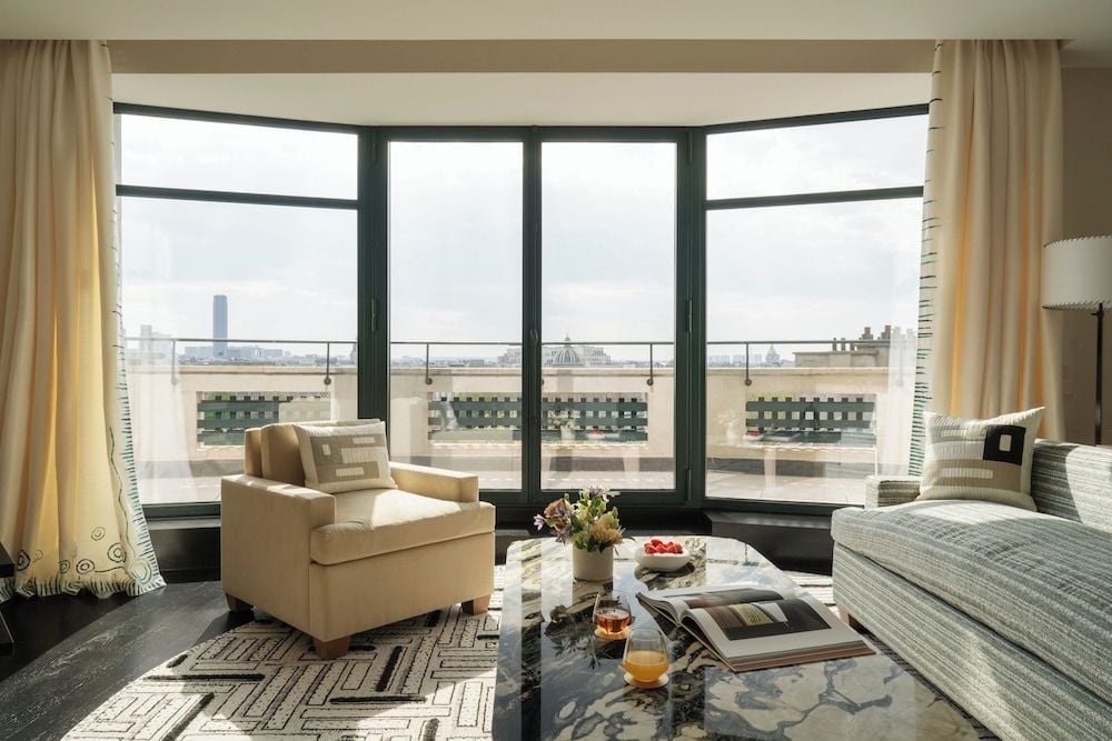 Floor-to-ceiling windows frame Parisian rooftops from this elegant suite, where honey-toned furnishings and geometric rugs create an intimate sanctuary above the bustling city.