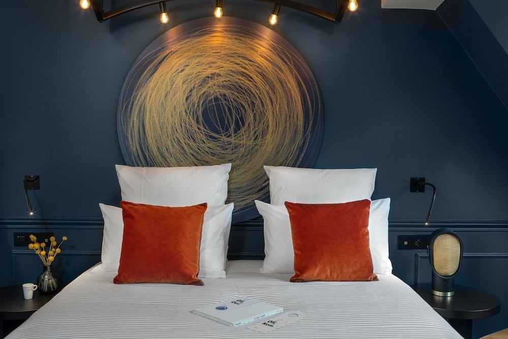 Warm Edison bulbs cast golden light over the sculptural headboard's swirling patterns, while burnt orange velvet pillows add richness to crisp white linens.