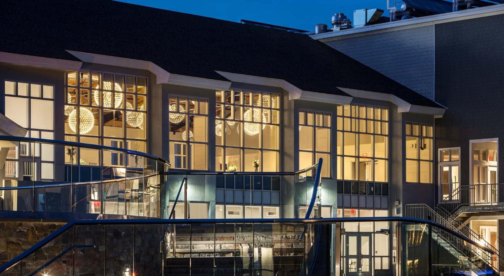 Floor-to-ceiling windows reveal warmly lit interiors where crystal chandeliers cast golden glows, while sleek railings frame expansive decks overlooking Maine's rugged coastline at dusk.