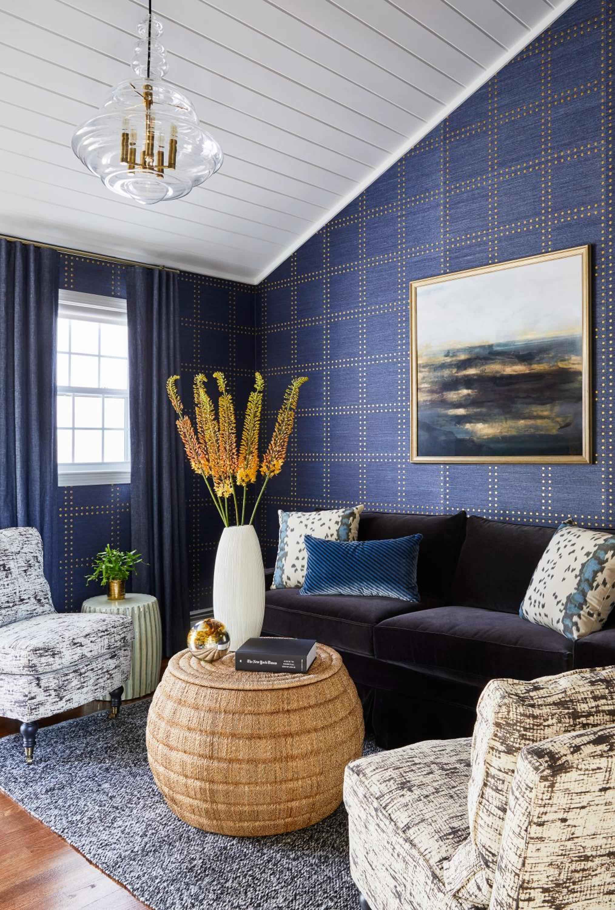 Navy grasscloth wallpaper with brass nail heads creates intimate drama in this sitting area, where wicker textures and coastal artwork evoke seaside sophistication.