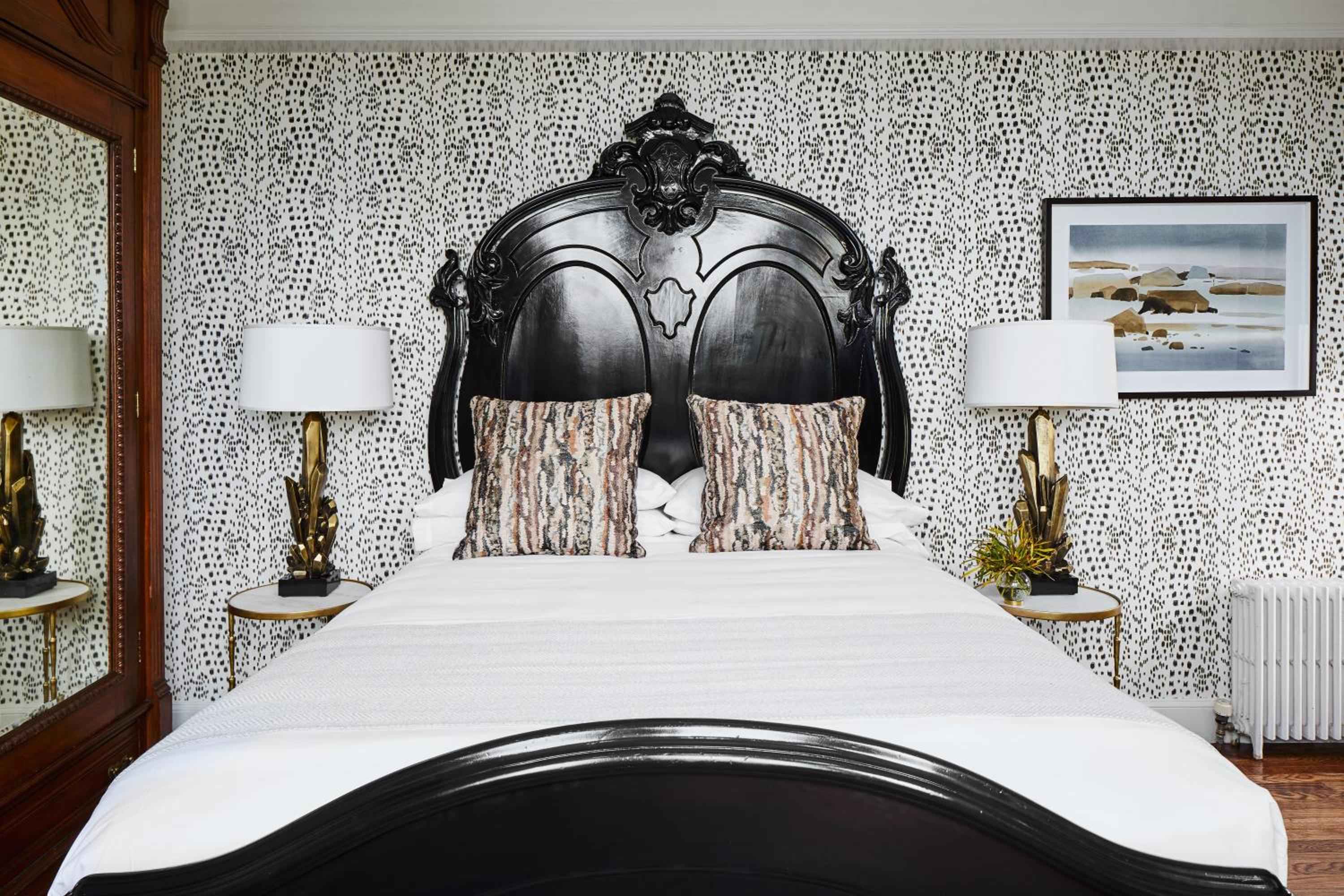 Ornate black headboard with baroque flourishes anchors crisp white linens against spotted wallpaper, where vintage glamour meets nautical New England charm in perfectly balanced contrast.