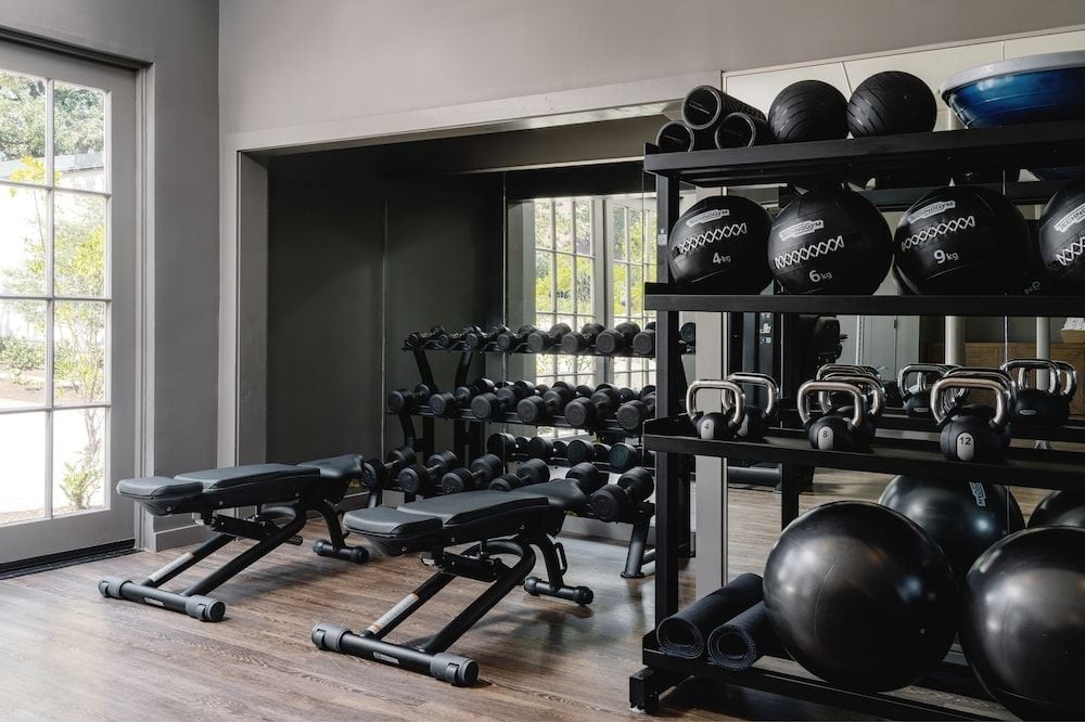 Natural light streams across polished weights and sleek equipment in this thoughtfully designed fitness sanctuary, where black steel meets warm wood floors beneath soaring ceilings.
