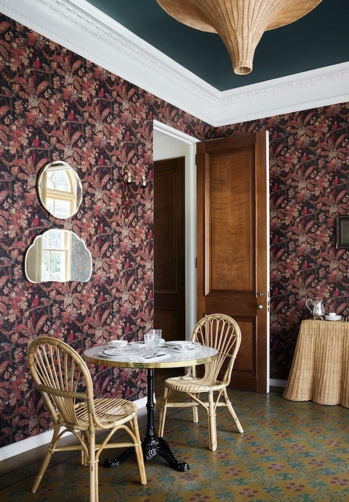 Moody floral wallpaper and rattan chairs create an intimate breakfast nook where dark botanical prints contrast beautifully with natural wicker textures and morning light.