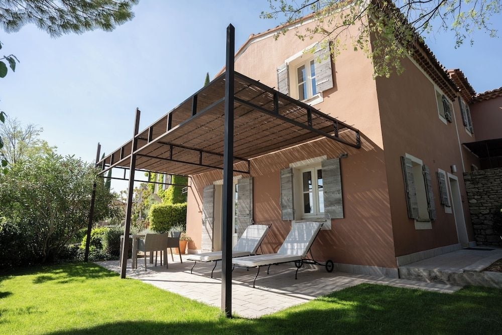 Weathered shutters frame terraced loungers beneath a rustic pergola, where dappled sunlight filters through reed screening onto manicured lawns and Provençal stone.